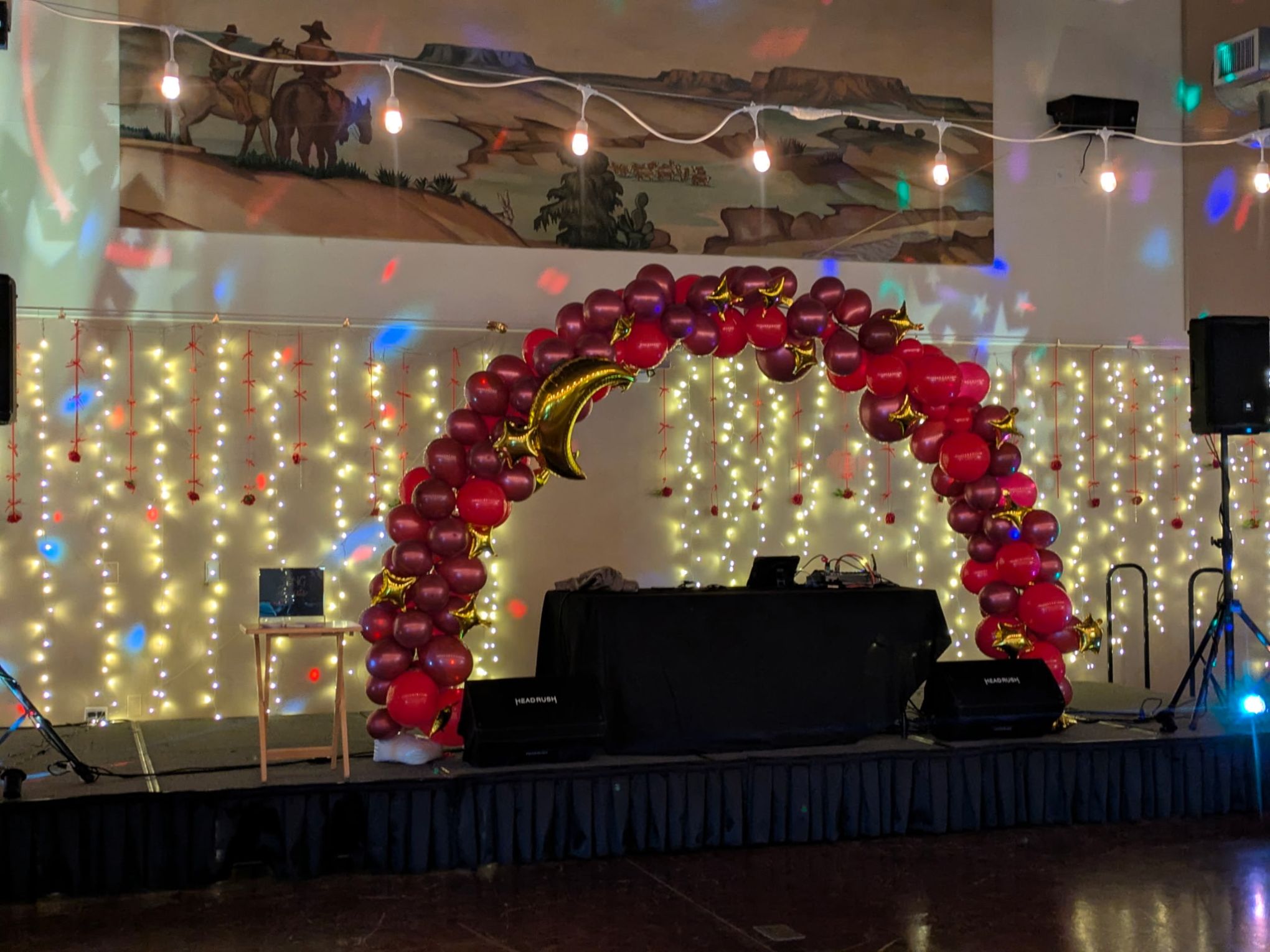 Stage with red balloon arch, twinkling lights, and DJ setup against a wall with hanging decorations.