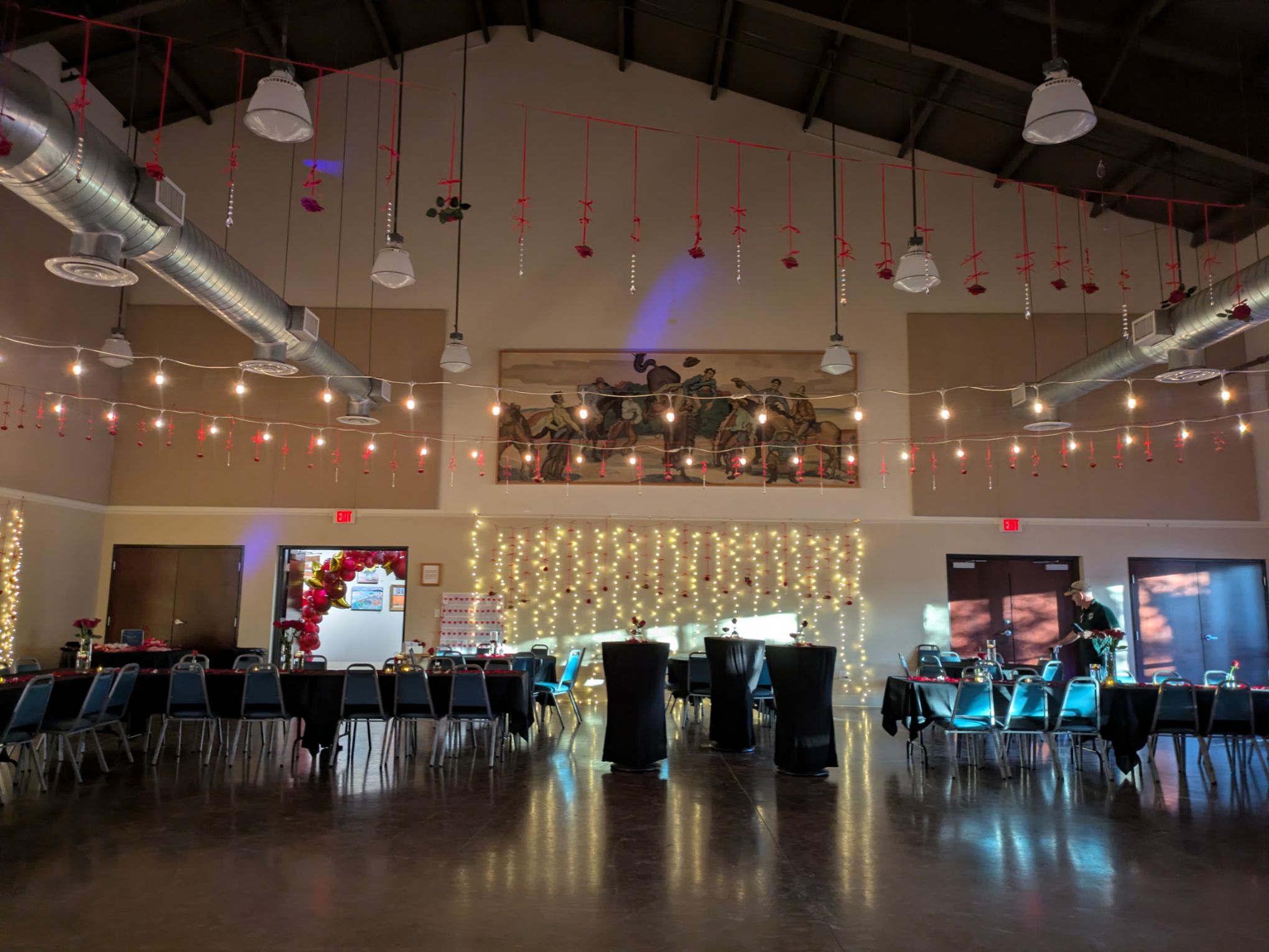 Decorated banquet hall with string lights, tables, chairs, and a mural on the wall.