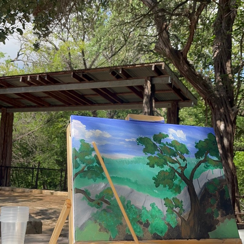 Person painting a landscape of trees and sky on a canvas in an outdoor setting.