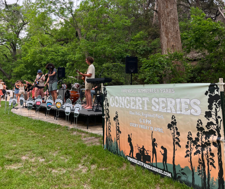 Outdoor band performing on a stage next to a banner for a concert series in a park setting.
