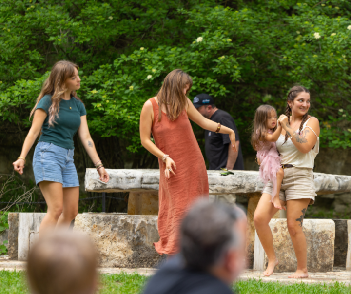 Four people dancing outdoors; one holding a child, with greenery in the background.