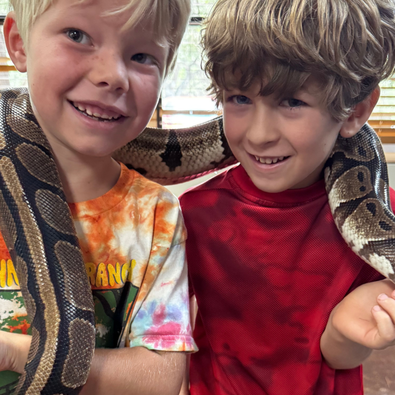Two children smiling with a snake draped around their shoulders indoors.