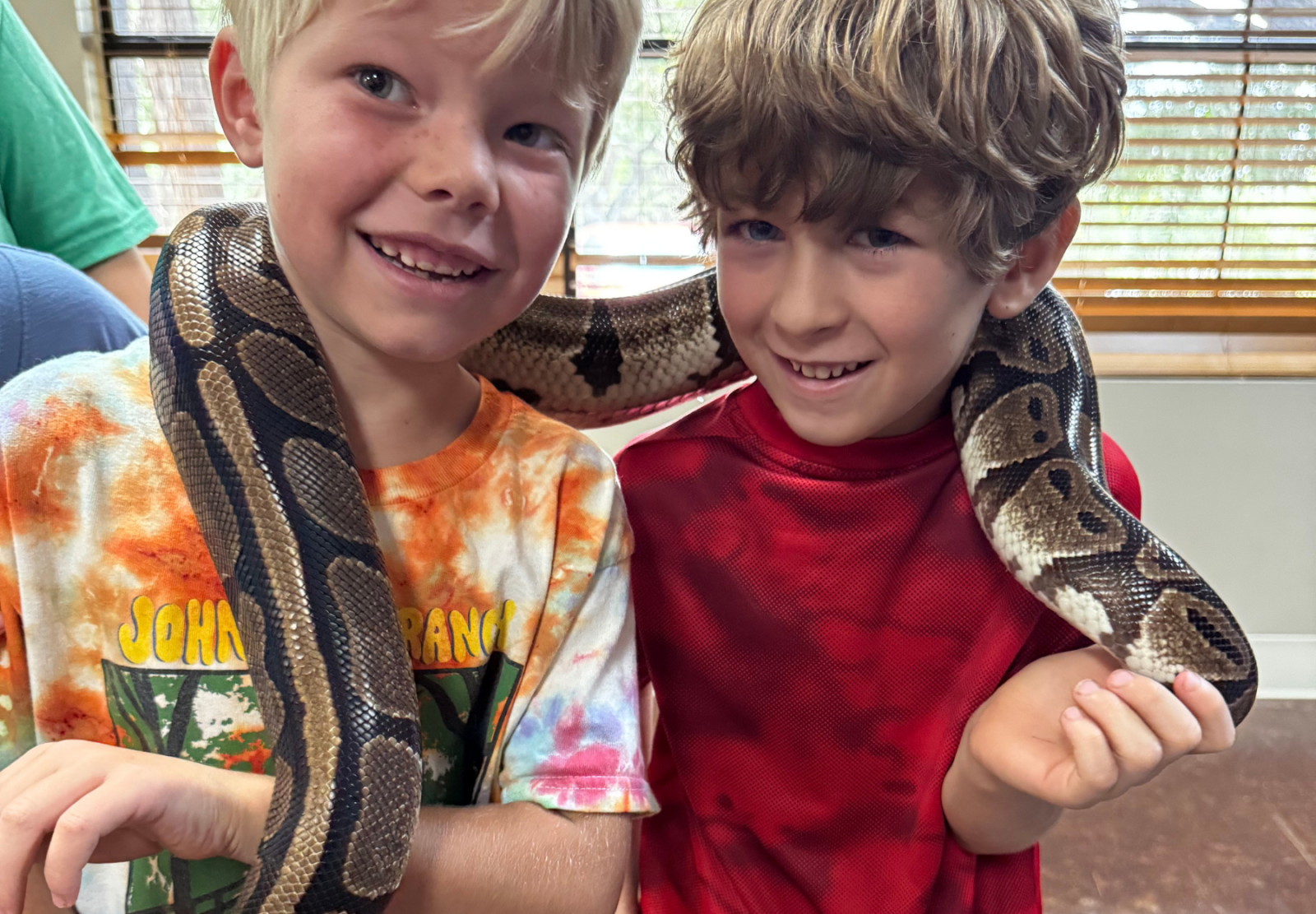 Two children smiling with a snake draped around their shoulders indoors.