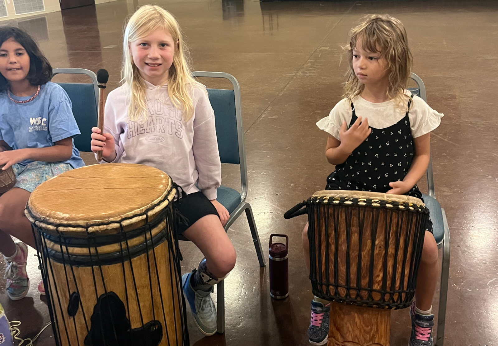 Three children sitting on chairs, each with a drum in front of them, smiling in a room with a wooden floor.