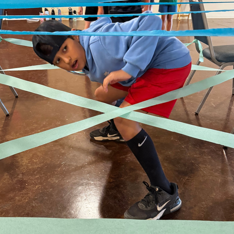 Child in cap and shorts navigates strings between chairs like a maze.