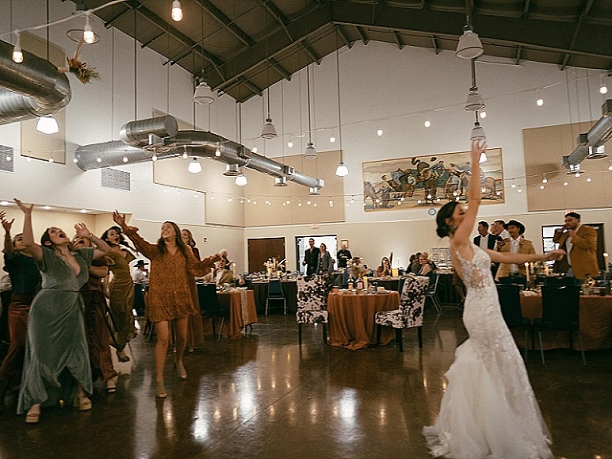 Bride tosses bouquet to women at indoor wedding reception.