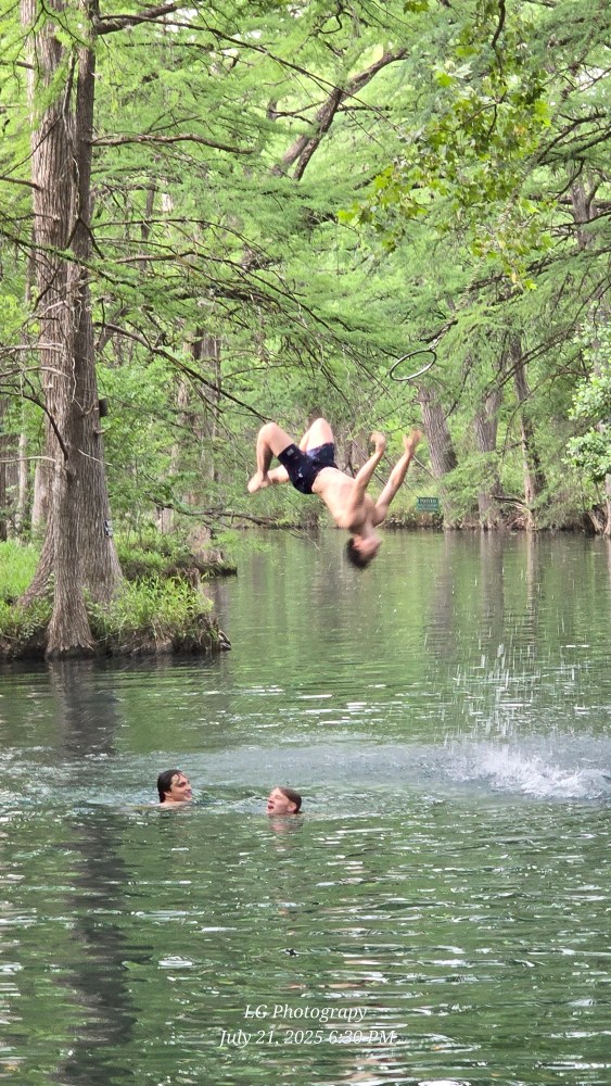 Person backflipping into a lake with two others swimming nearby, surrounded by trees.