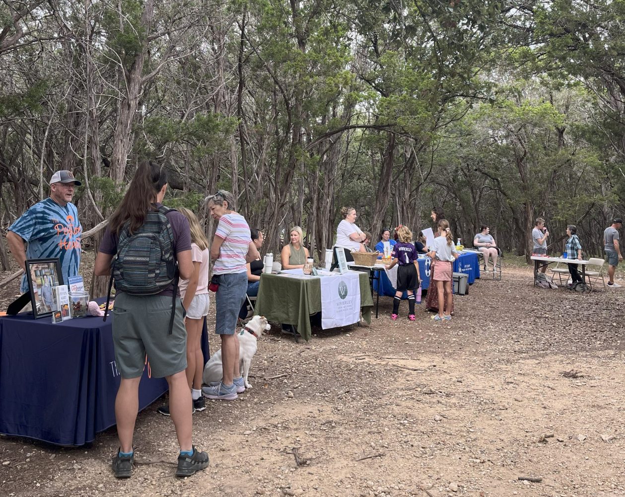 People gathered at outdoor tables in a wooded area under a cloudy sky.