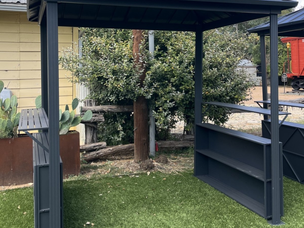 Black metal gazebo with shelves on green grass, wooden wall and plants in background.