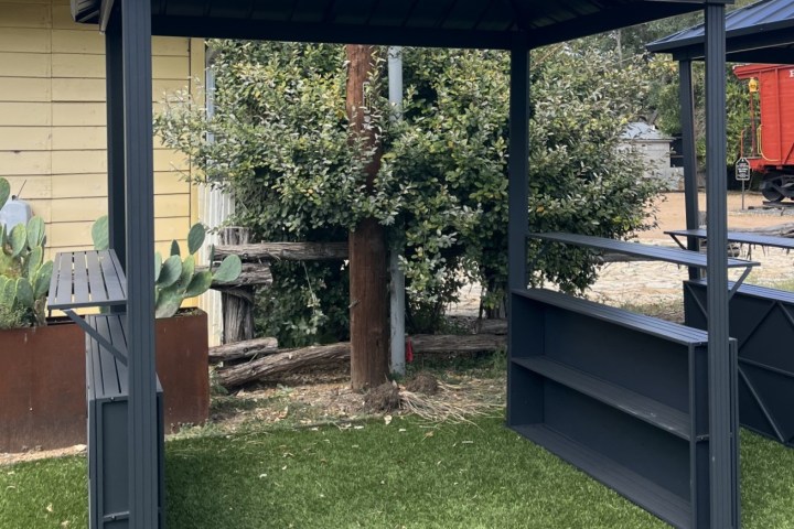 Black metal gazebo with shelves on green grass, wooden wall and plants in background.