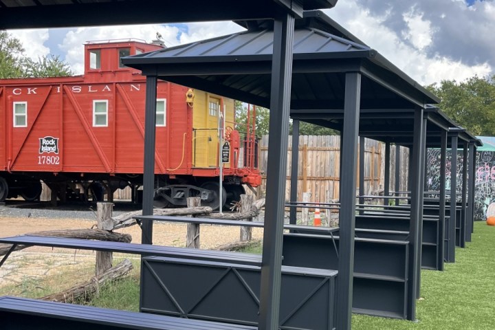 Outdoor seating area with metal structures beside a red vintage train on green grass under a blue sky.