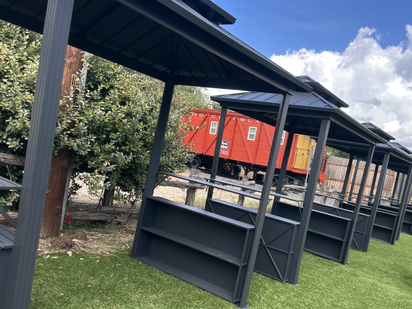 Black gazebos on green grass with a red train car and bushes in the background.