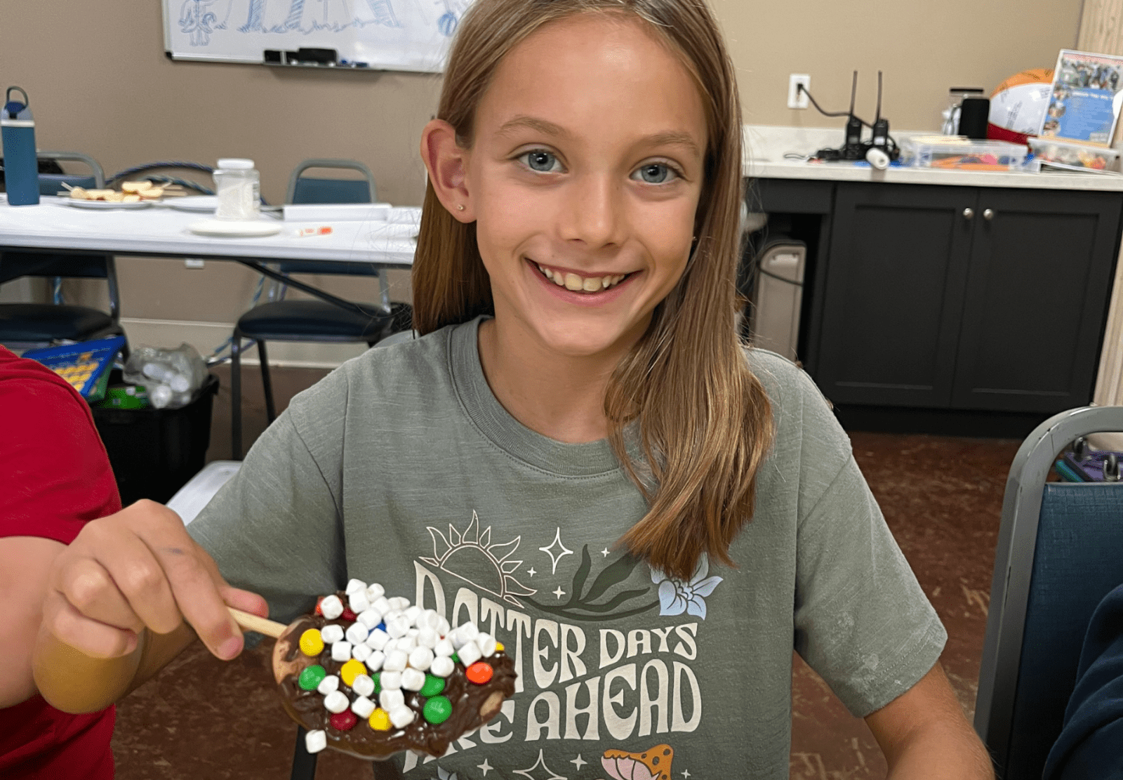 Girl smiling, holding a plate with marshmallows and candy in a room with chairs and a cabinet in the background.