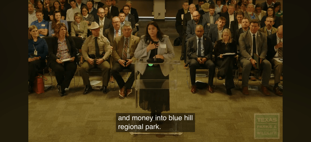 Woman speaks at podium in crowded meeting; audience seated in rows.