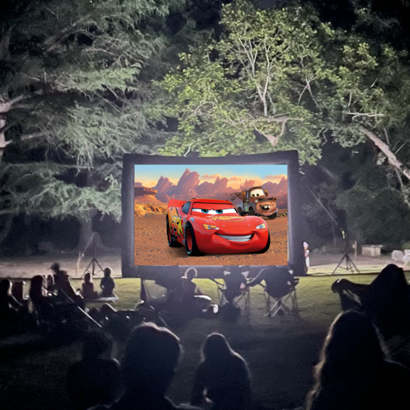 Outdoor movie night with people watching 'Cars' on a screen under trees.