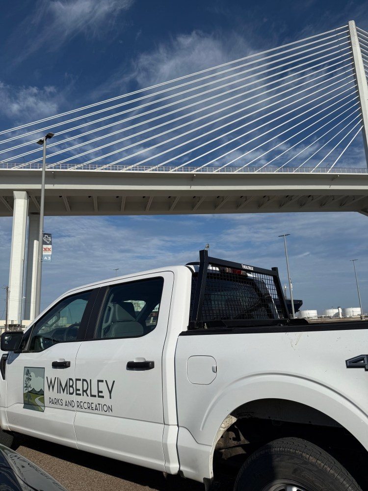 White truck with 'Wimberley Parks and Recreation' logo under a cable-stayed bridge.