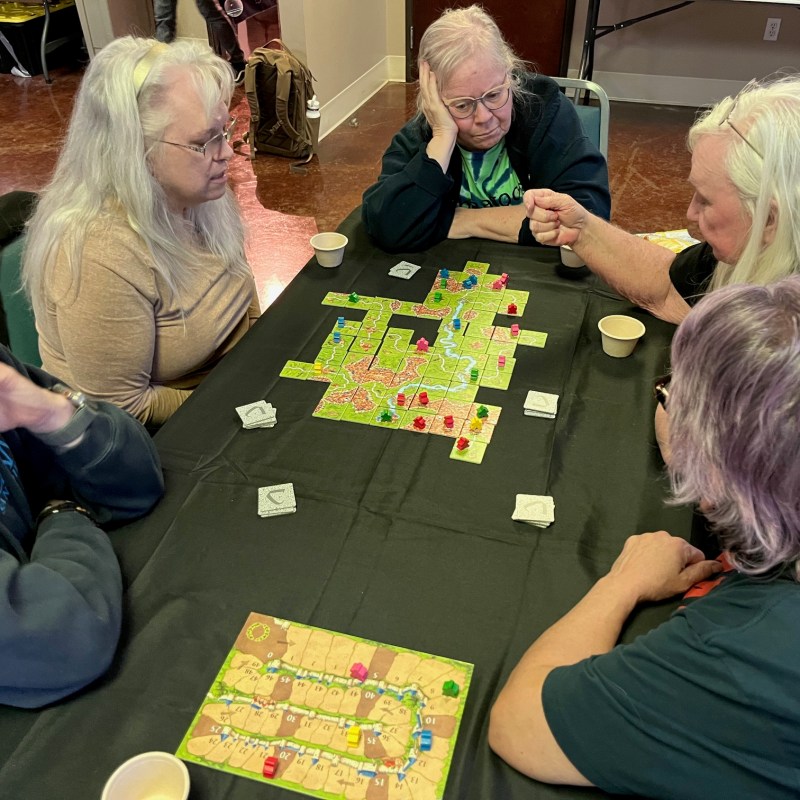 Five people playing a board game with colorful tiles and pieces on a table.