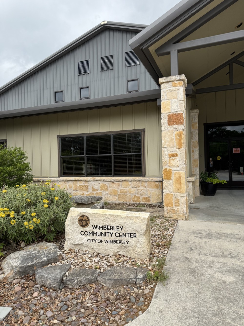 Exterior of Wimberley Community Center with stone sign and yellow flowers.