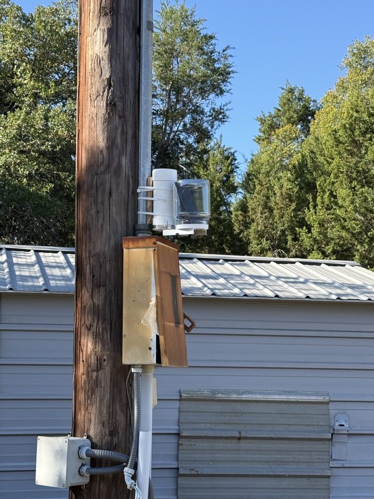 Wooden utility pole with equipment, shed and trees in background under clear sky.