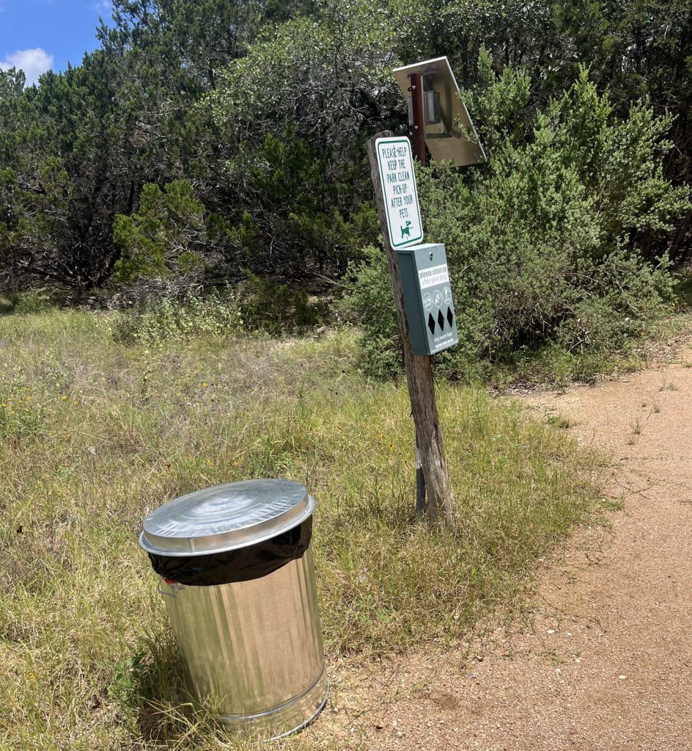 Trail sign with bag dispenser and trash can on a dirt path.