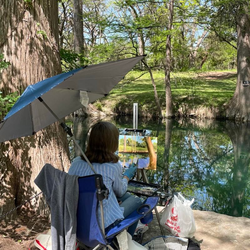 Person painting by a riverside under an umbrella, surrounded by trees.