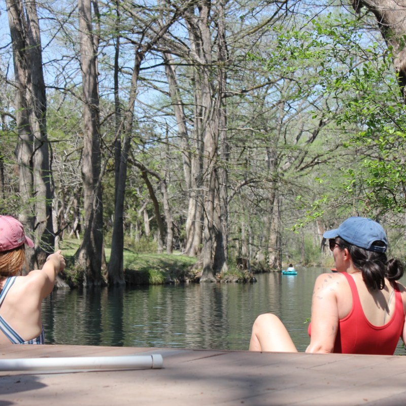 Two women sitting by a wooded river, one pointing, both in swimwear.