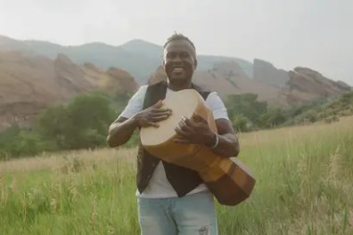 Person holding guitar in grassy field with rocky hills in background.