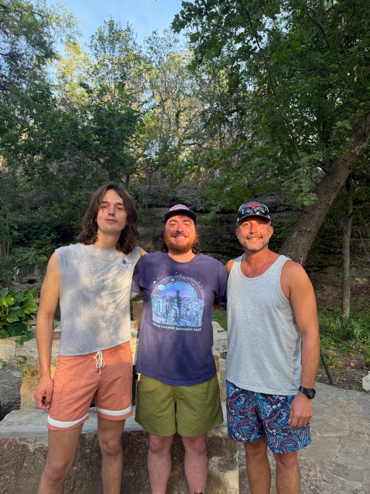 Three people smiling outdoors in a wooded area with trees in the background.