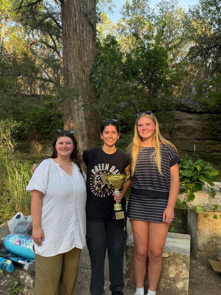 Three people outdoors, one holding a trophy, surrounded by trees and greenery.