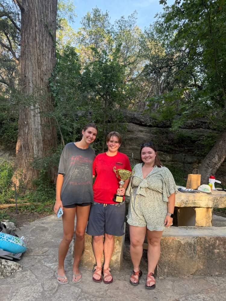 Three people posing outdoors with a trophy in a wooded area.