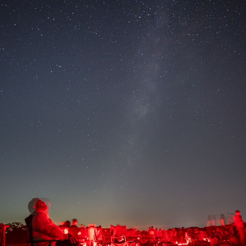 Starry night sky with people sitting, red lights on ground.