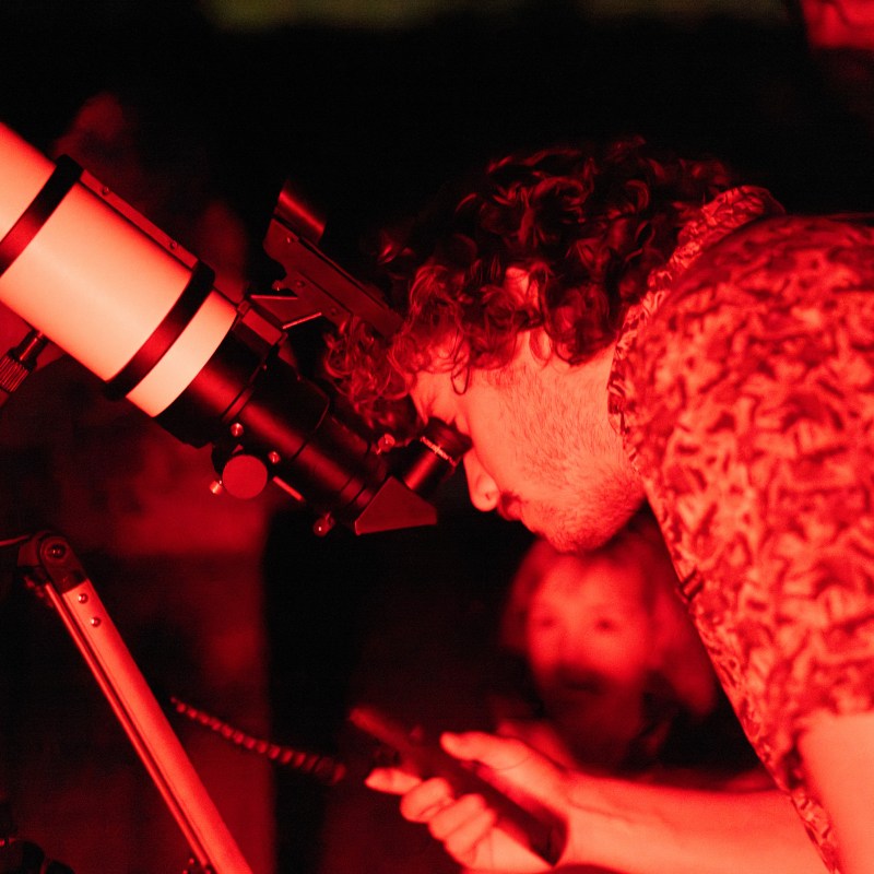 Person observing stars through a telescope at night, illuminated by red light.