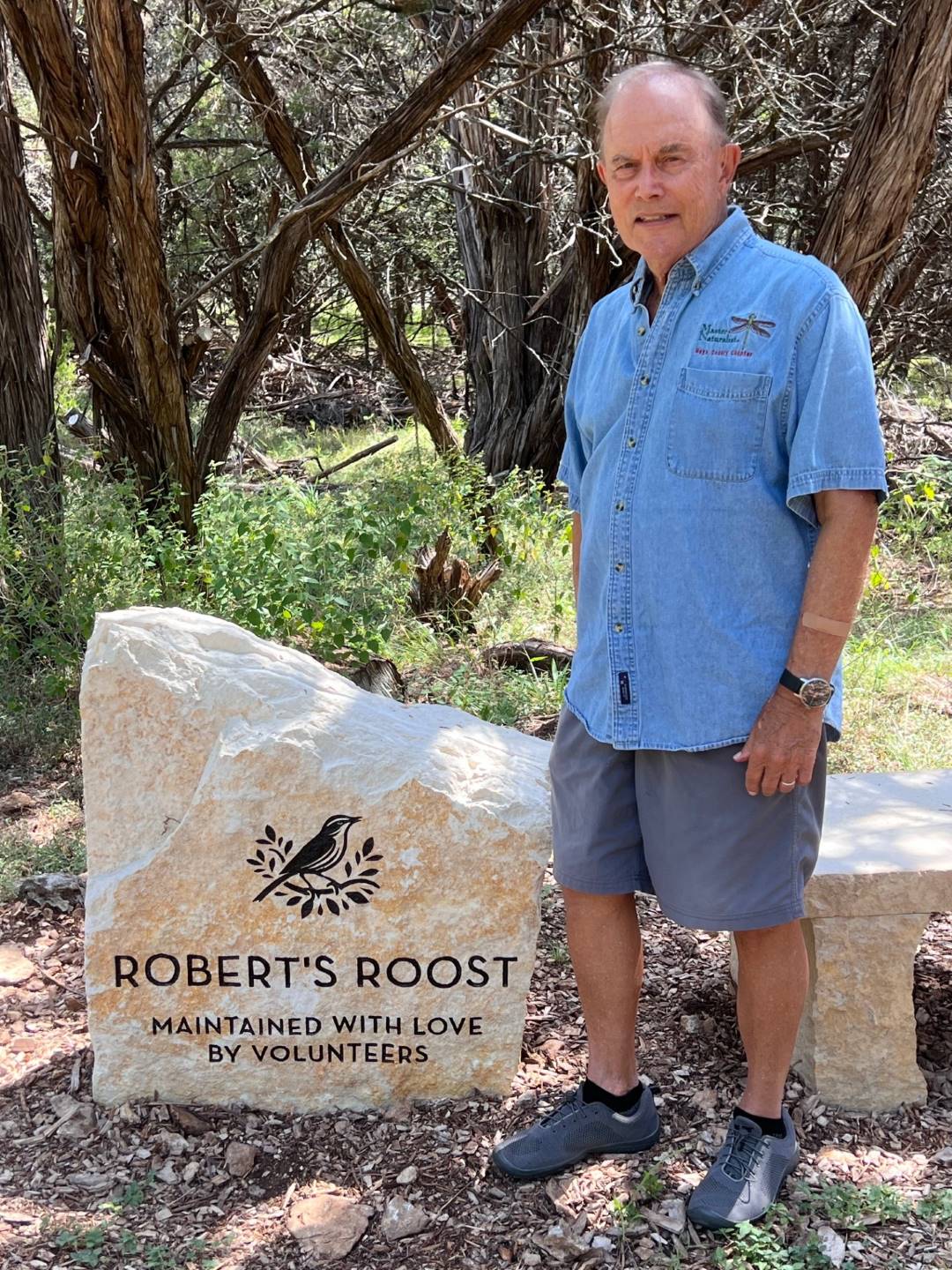 Man standing beside a stone sign reading 'Robert's Roost' in a wooded area.