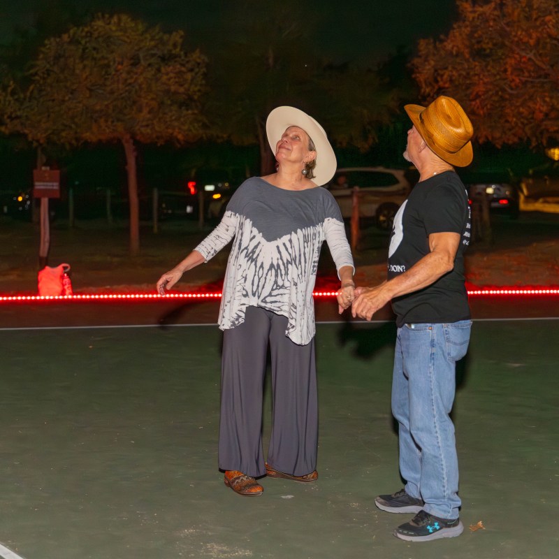 Two people wearing hats dance on an outdoor court at night.