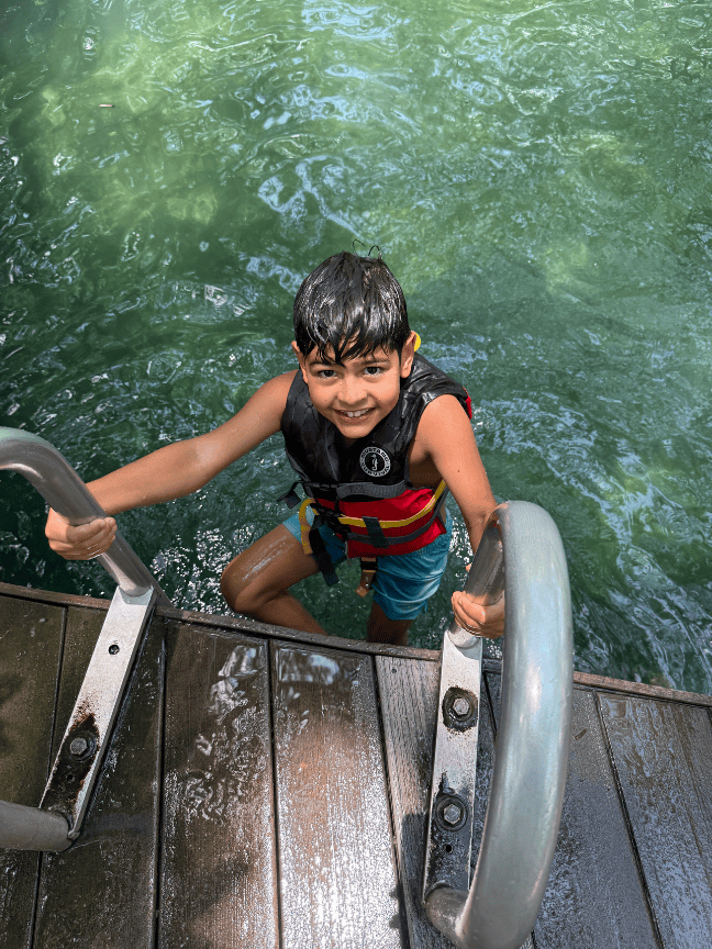 Child in a life jacket climbing a ladder from green water onto a wooden dock.
