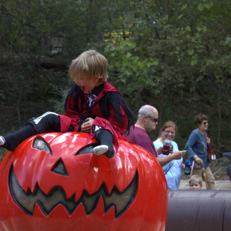 Child in costume on inflatable jack-o'-lantern outdoors with people around.