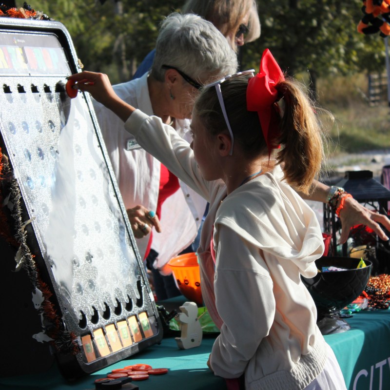 Child playing a Plinko game at a festival booth, with adults assisting.