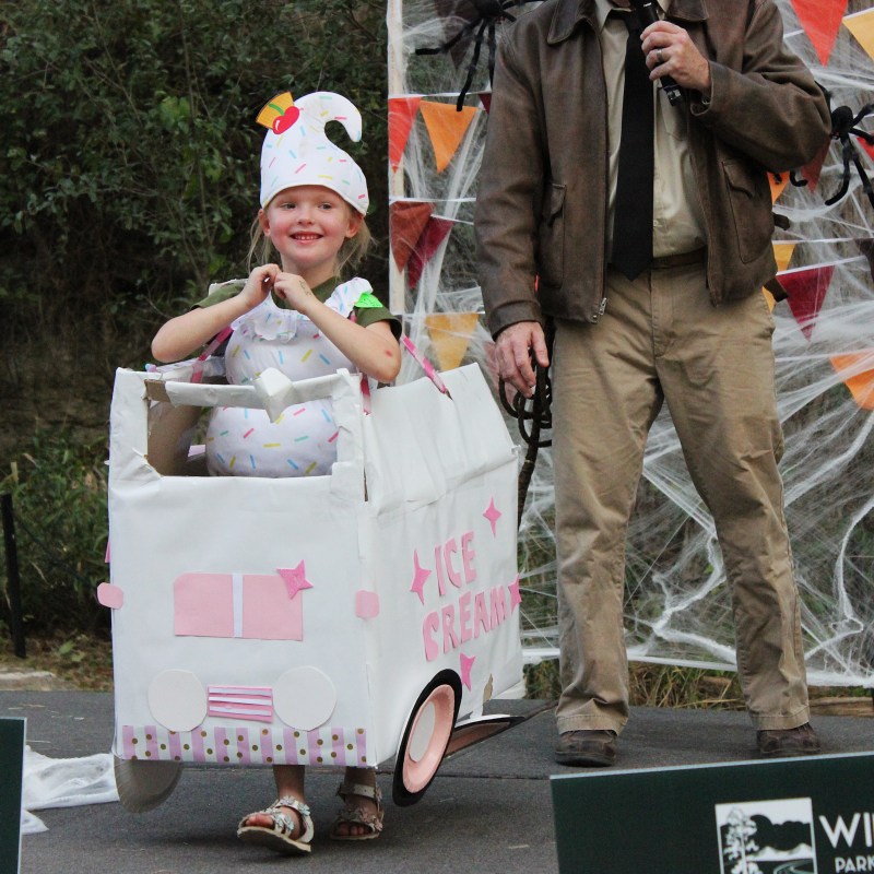 Child in ice cream truck costume, smiling on stage with adult nearby.